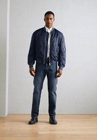 Marine bomberjakke med ribbede mansjetter og nederkant, iført over en hvit skjorte, sammen med mørke denimjeans og svarte støvler.