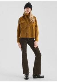 Vrouw in een mosterdkleurig corduroy overhemd, bruine corduroy broek, zwarte beanie en zwarte enkellaarzen, staand tegen een effen achtergrond.