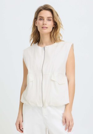 Femme aux cheveux blonds portant un haut blanc sans manches avec une fermeture éclair à l'avant et des poches, ainsi qu'un pantalon blanc, sur un fond clair uni.