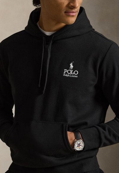 Férfi fekete Polo Ralph Lauren kapucnis pulóverben, fehér logóval, kezét zsebében tartja, és fehér számlapos, fekete bőrszíjas karórát visel.