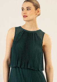 Smaragdgrünes, gefaltetes Kleid mit ärmellosem Design, das einen runden Ausschnitt und eine geschichtete Textur aufweist, die eine fließende Silhouette kreiert.