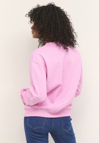 Mujer con cabello rizado, vistiendo un suéter rosa y jeans azules, mirando hacia la izquierda contra un fondo claro y liso.