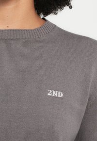 Maglione grigio chiaro con scollo a coste. Presenta testo ricamato bianco "2ND" sul davanti. Il materiale sembra morbido e strutturato.