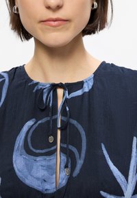 Blouse bleu marine avec un col à nouer, motifs abstraits bleus et tissu texturé. Détails remarquables : accents en métal circulaire.