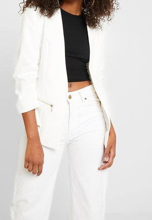 Vrouw die een witte blazer draagt over een zwart cropped topje en witte, hooggetailleerde broek, staand tegen een effen achtergrond.