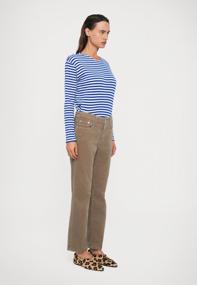 Vrouw die in profiel staat, gekleed in een blauw-wit gestreepte langemouwen shirt, beige corduroy broek en luipaardprint instapschoenen.