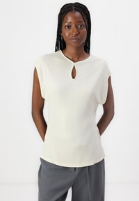 Haut sans manches de couleur beige clair avec un décolleté en forme de trou de serrure, en tissu lisse et coupe décontractée, associé à un pantalon gris. Design simple et minimaliste.