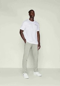Manlig modell som bär en vit Champion t-shirt, ljusgråa casual byxor och vita sneakers, står med en hand i fickan mot en enfärgad bakgrund.