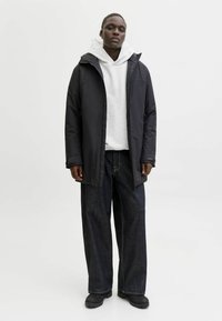 Czarna wodoodporna kurtka, szara bluza z kapturem, szerokie ciemnoniebieskie dżinsy i czarne buty. Gładki materiał, luźny krój, minimalistyczny design.