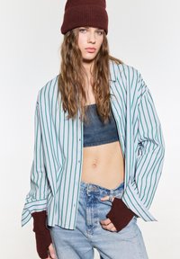 Pruhovaná košile na knoflíky v světle modré barvě se zelenými pruhy, vrstvená přes tmavý crop top, v kombinaci s džínovými kalhotami s vysokým pasem a hnědými rukavicemi s ohrnutými manžetami.