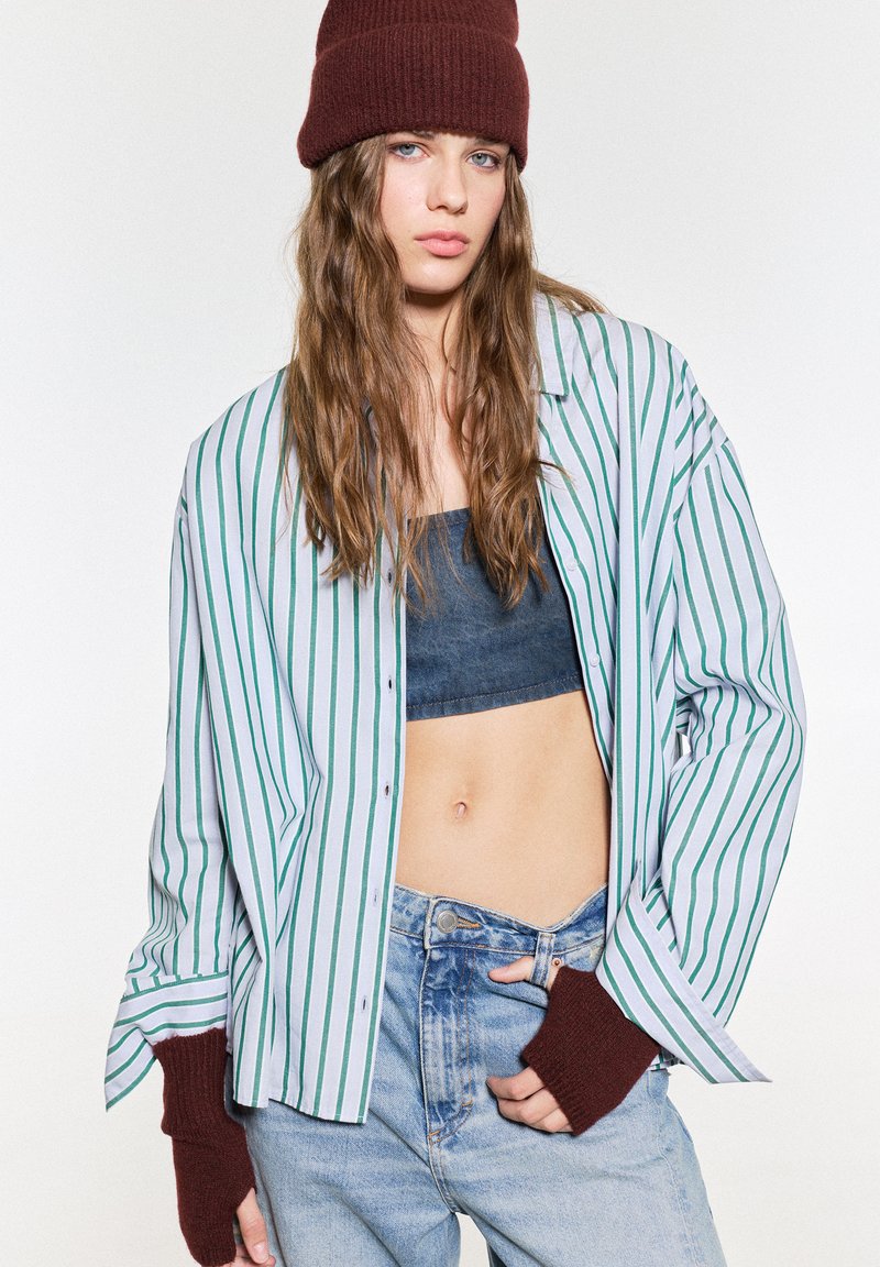 Pruhovaná košile na knoflíky v světle modré barvě se zelenými pruhy, vrstvená přes tmavý crop top, v kombinaci s džínovými kalhotami s vysokým pasem a hnědými rukavicemi s ohrnutými manžetami.