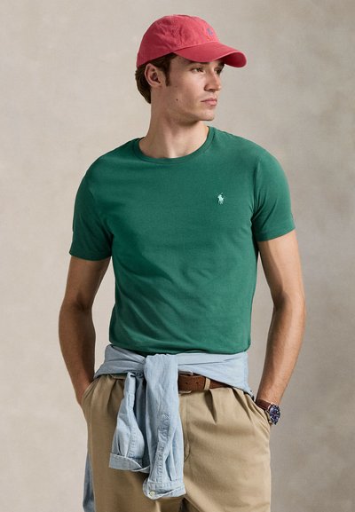 T-shirt vert à manches courtes avec un col rond et un petit logo brodé. Porté avec un short kaki et une chemise bleu clair nouée autour de la taille.