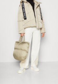 Beige dunjakke med hætte og sorte logostropper, båret over en sort turtleneck, kombineret med hvide bukser og en matchende beige tote taske.