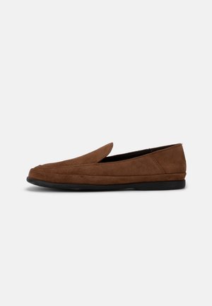Mocassin en daim marron avec semelle noire basse et design sans lacets, présenté en profil sur un fond gris clair uni.