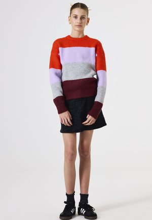 Pull en tricot avec des rayures horizontales orange, lavande, gris et bordeaux. Encolure ras du cou et poignets côtelés. Associé à une jupe noire.