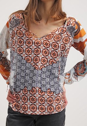 Woman wearing a long-sleeve V-neck blouse with orange, black, and white floral and geometric patterns, paired with black pants.