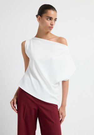 Donna che indossa una blusa bianca a spalle scoperte e pantaloni bordeaux a vita alta, in piedi davanti a uno sfondo bianco uniforme.