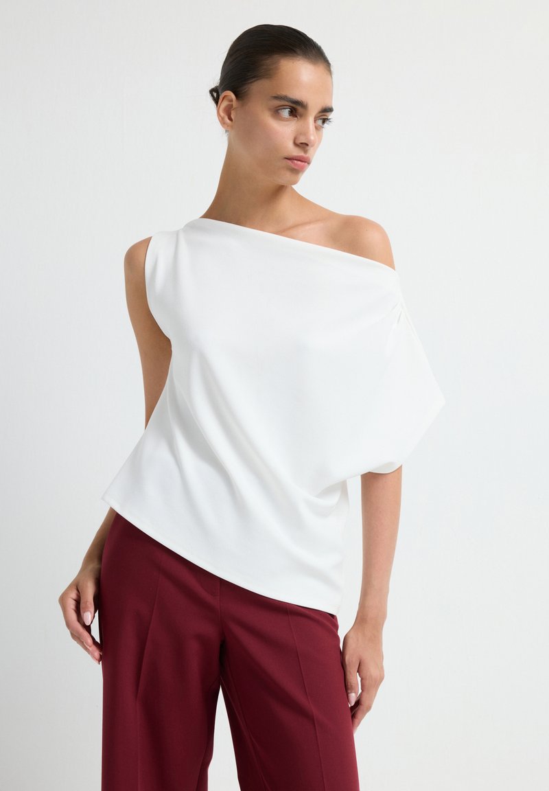 Donna che indossa una blusa bianca a spalle scoperte e pantaloni bordeaux a vita alta, in piedi davanti a uno sfondo bianco uniforme.