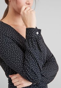 Blouse noire à pois avec un décolleté en V profond et des manches longues. Les manches sont dotées de poignets boutonnés et d'une texture froncée au niveau des poignets.