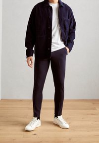 Navy corduroy jacket with button closure and two pockets, paired with fitted navy pants and white sneakers on a wooden floor.