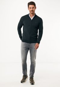 Suéter de cremallera en verde oscuro con un diseño ajustado, combinado con jeans gris claro y zapatillas oscuras. Textura suave, con un mínimo de marca en el suéter.