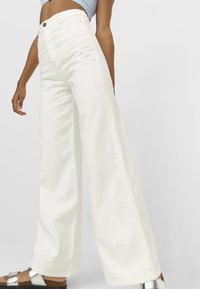 Pantalons en denim blancs à jambe large avec une taille haute, dotés d'une fermeture à bouton et de détails sur les coutures latérales, associés à des sandales métalliques.