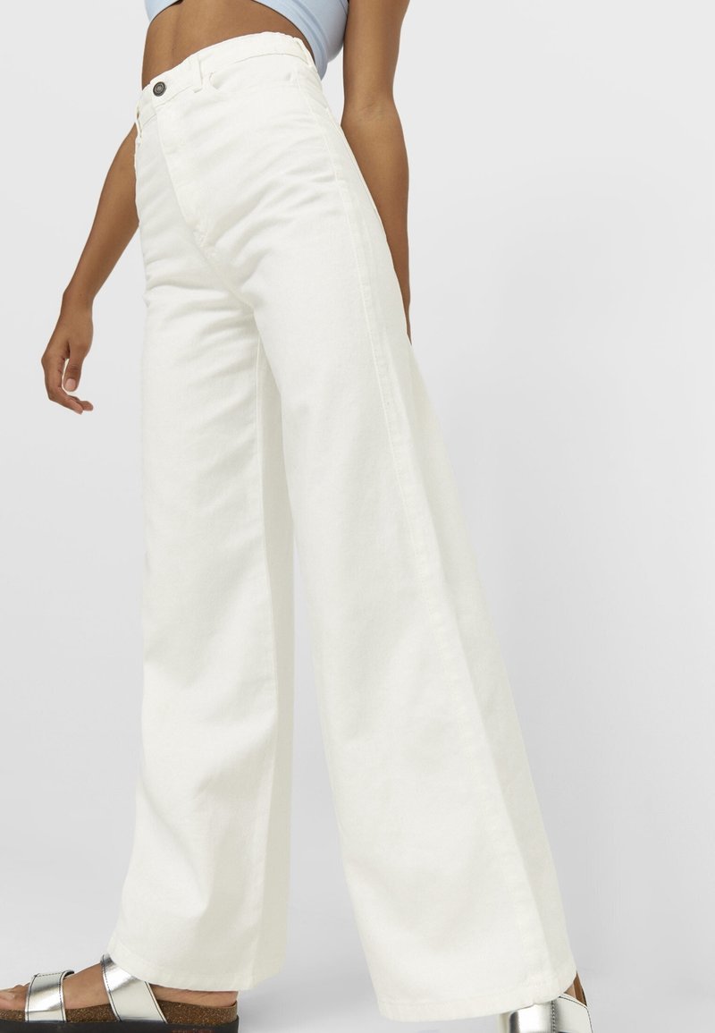 Pantalons en denim blancs à jambe large avec une taille haute, dotés d'une fermeture à bouton et de détails sur les coutures latérales, associés à des sandales métalliques.