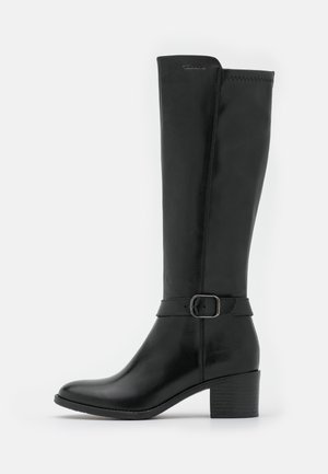 Botte haute en cuir noir avec un petit talon bloc et une sangle décorative avec une boucle argentée autour de la cheville.