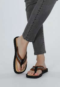 Brown woven flip-flops with black straps, featuring a textured sole and decorative accents. Worn with gray ankle-length jeans.