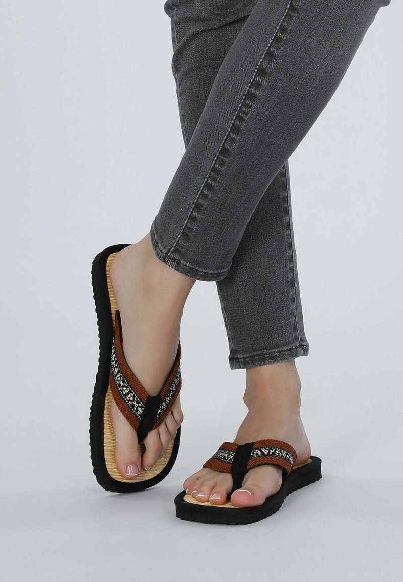 Brown woven flip-flops with black straps, featuring a textured sole and decorative accents. Worn with gray ankle-length jeans.