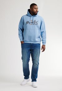 Světle modrá mikina s kapucí a černým logem, v kombinaci se středně zesvětlými džíny a bílými teniskami. Jasný, neformální sportovní styl.