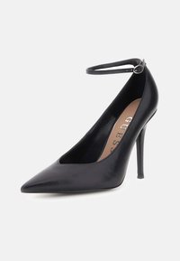 Escarpin noir en cuir verni à talon haut avec bout pointu et bride de cheville, présentant un design épuré et une texture brillante. Nom de la marque visible à l'intérieur.