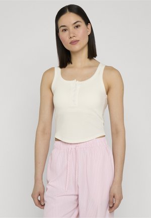 Ung kvinde med glat mørkt hår iført en hvid ærmeløs knaplukket crop top og lyserøde, stribede bukser med snøre, stående mod en ensfarvet baggrund.