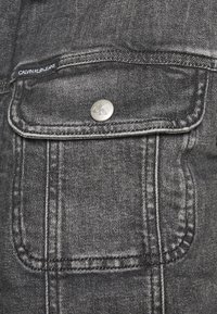 Jeans Calvin Klein en denim gris oscuro con un bolsillo frontal con cierre a presión metálico y detalle de costura visible.