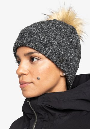 Strikket grå beanie med en faux fur pom-pom, der har et tekstureret design og en subtil logoaccent, rammet ind mod en sort jakke.