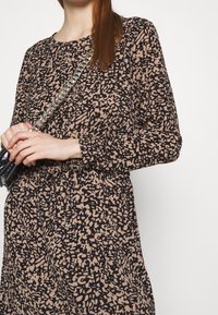 Långärmad klänning i svart och taupe abstrakt mönster, draperad vid midjan. Modellen håller en liten svart handväska med en silverkedjesnor.