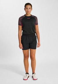 Svart sporttopp med rosa mönstrade ärmar, kombinerad med svarta shorts. Materialet verkar vara lättviktigt; inkluderar logodetaljer på tröjan och skorna.