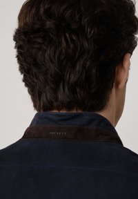 Mornarica suđena košulja s tamnosmešnim ovratnikom, na kojem je utisnut naziv brenda "HACKETT" na leđima ovratnika. Glatka tekstura i klasičan dizajn.