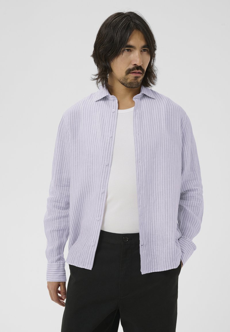 Homme aux cheveux brun moyens portant une chemise à rayures violettes claires boutonnée sur un débardeur blanc et un pantalon noir, les mains dans les poches.