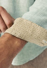Puño de camisa de patrón verde con un puño beige en contraste que presenta un diseño geométrico blanco, revelando detalles de tejido texturizado.