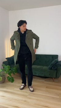 Jonge man past een olijfgroene jas aan over een zwarte hoodie, staand op een houten vloer in een woonkamer met een donkergroene fluwelen bank en een potplant.