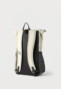 Μπεζ και μαύρο σακίδιο πλάτης με ρολάρι κορυφής, επενδεδυμένους ιμάντες, πλέγμα πίσω πάνελ, πλαινές τσέπες από πλέγμα και χειρολαβή στην κορυφή σε λευκό φόντο.