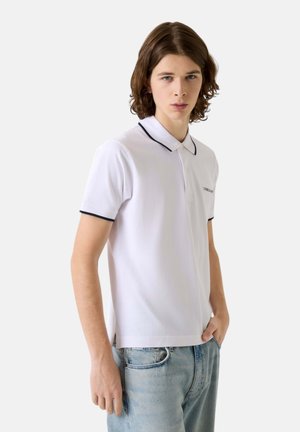 Giovane uomo con capelli di media lunghezza che indossa una polo bianca con rifiniture nere e jeans azzurri, in piedi con una mano in tasca.