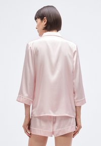Ροζ σατέν σετ πιτζάμες που περιλαμβάνει πουκάμισο με κουμπιά και μανίκια τριών τετάρτων και σορτς, με απαλή, γυαλιστερή υφή και αντίθετες λεπτομέρειες.