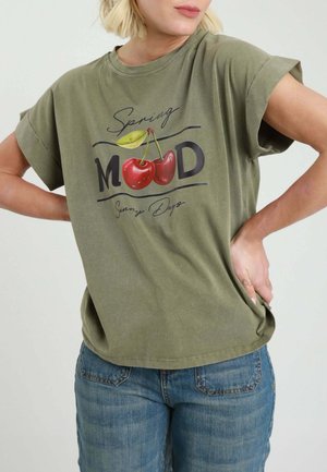 Persona che indossa una maglietta verde oliva con maniche arrotolate e grafica di ciliegie, abbinata a jeans blu, con le mani posate sui fianchi.