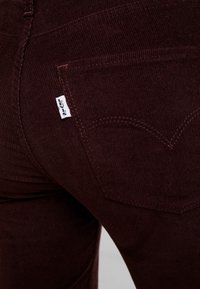 Jeans de pana burdeos con un bolsillo trasero que lleva una etiqueta blanca de Levi's y un patrón de costura distintivo. Tela texturizada visible.