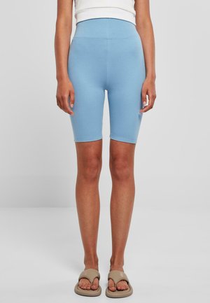 Kvinde iført lyseblå cykelshorts med højtaljet og beige slip-on sandaler, stående mod en ensfarvet lys baggrund.