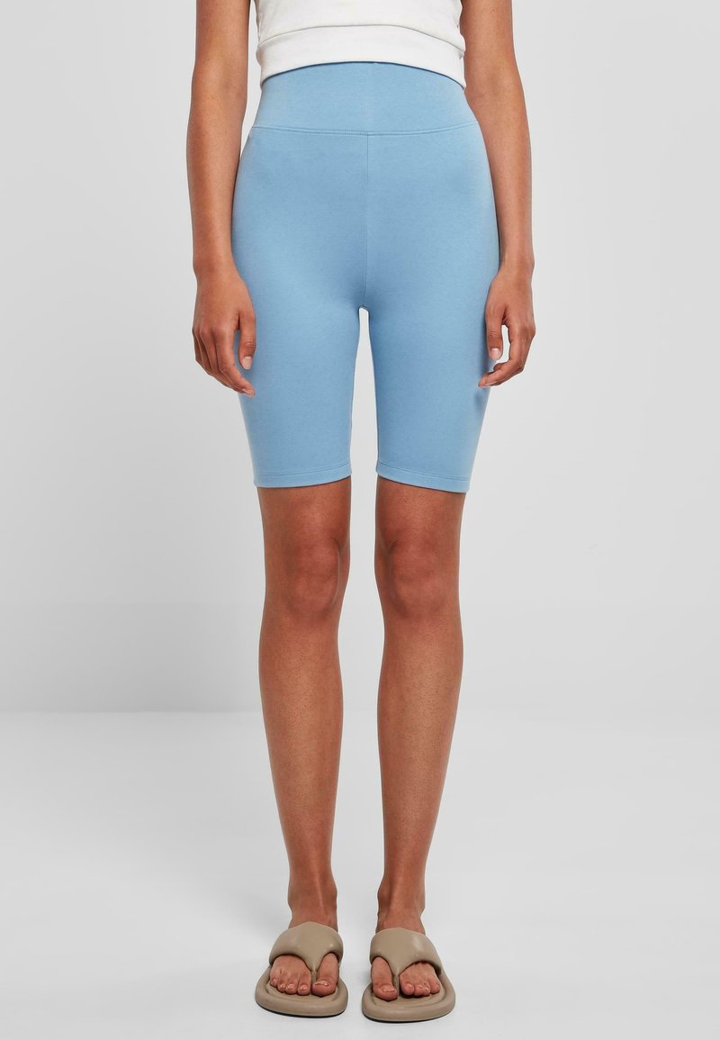 Femme portant un short cycliste bleu clair taille haute et des sandales beige à enfiler, debout devant un fond clair uni.