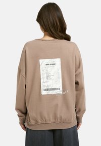 Frau trägt einen braunen Sweatshirt mit einem weißen aufgedruckten Beleg-Graphic und dem Text "Freundliche Erinnerung" auf dem Rücken, kombiniert mit dunklen Hosen.