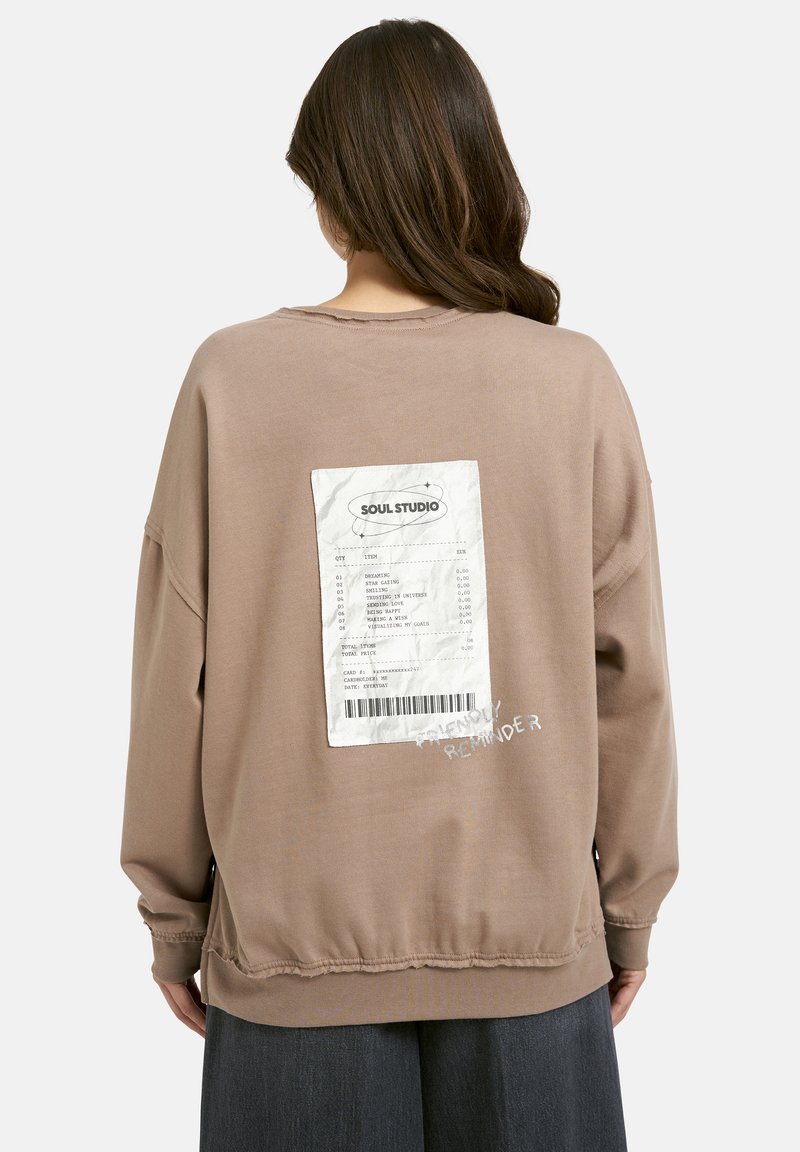 Frau trägt einen braunen Sweatshirt mit einem weißen aufgedruckten Beleg-Graphic und dem Text "Freundliche Erinnerung" auf dem Rücken, kombiniert mit dunklen Hosen.